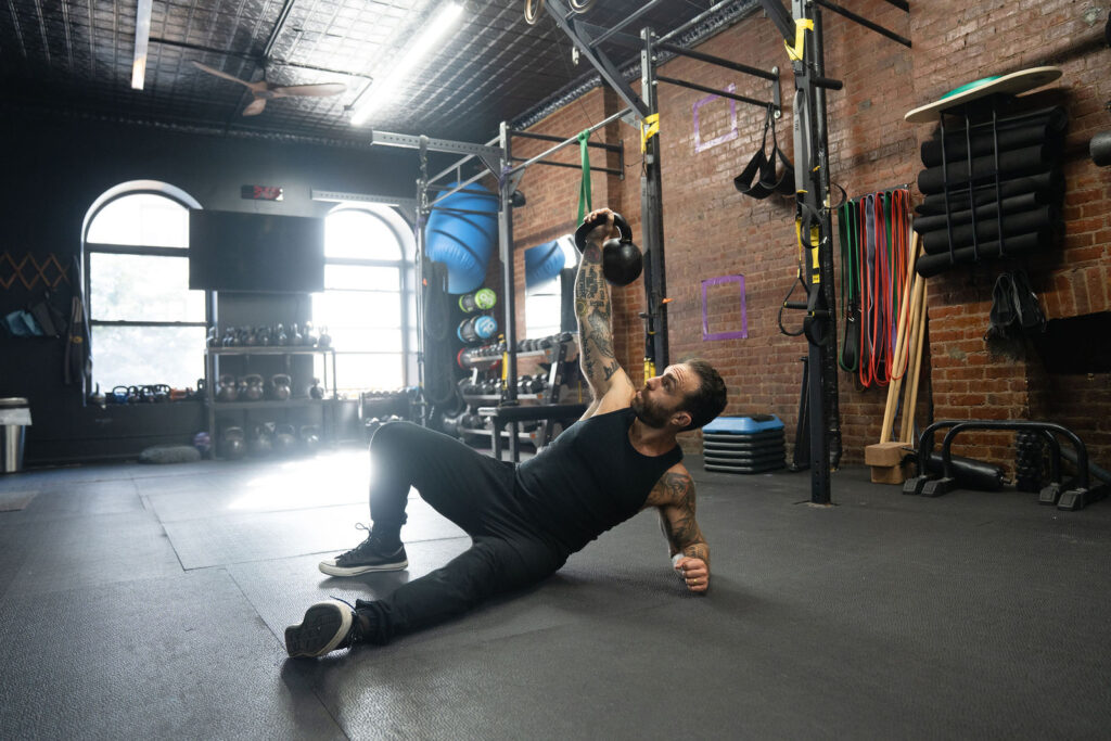 Dave Cash performing a kettlebell get-up on the gym floor in window light